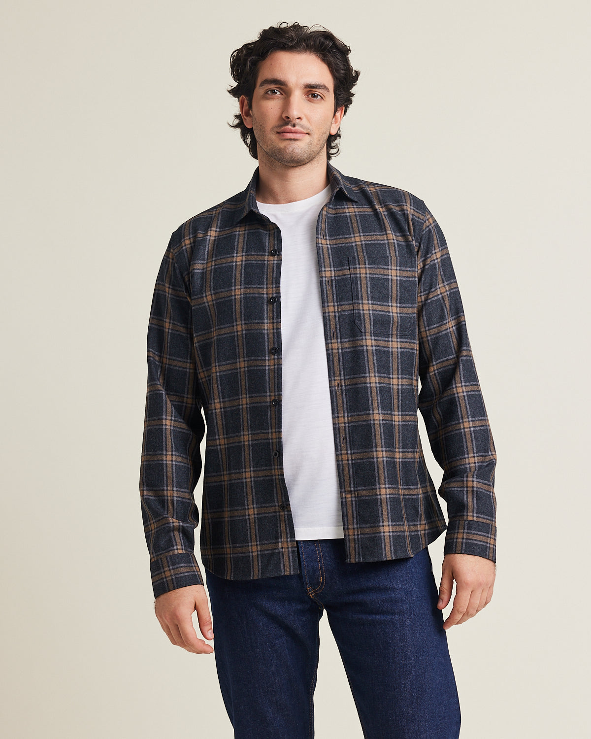 Man wears a merino wool flannel shirt in dark grey brown plaid with chestpocket open over a white merino wool T-shirt on a dark denim jeans