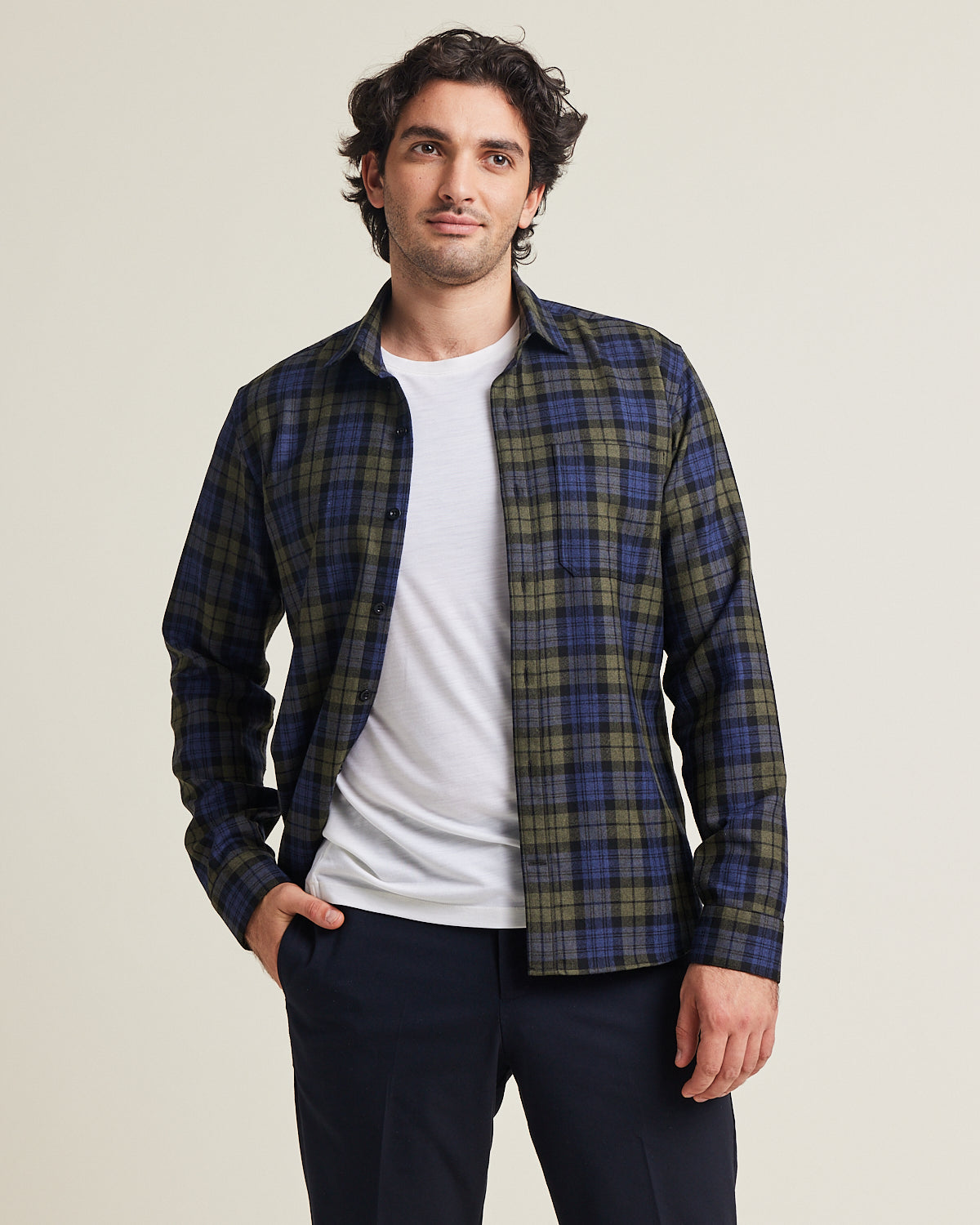 Man wears a merino flannel shirt in plaid blue green color with chestpocket open over a white merino wool T-shirt on a dark navy pants