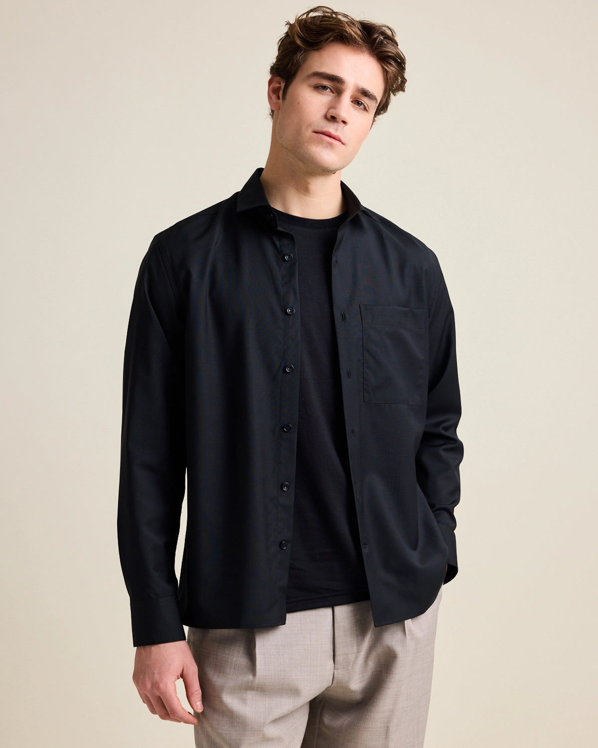 Man wears a black merino wool shirt open over a black merino wool T-shirt from Wolk. The shirt has a relaxed, wide fit.