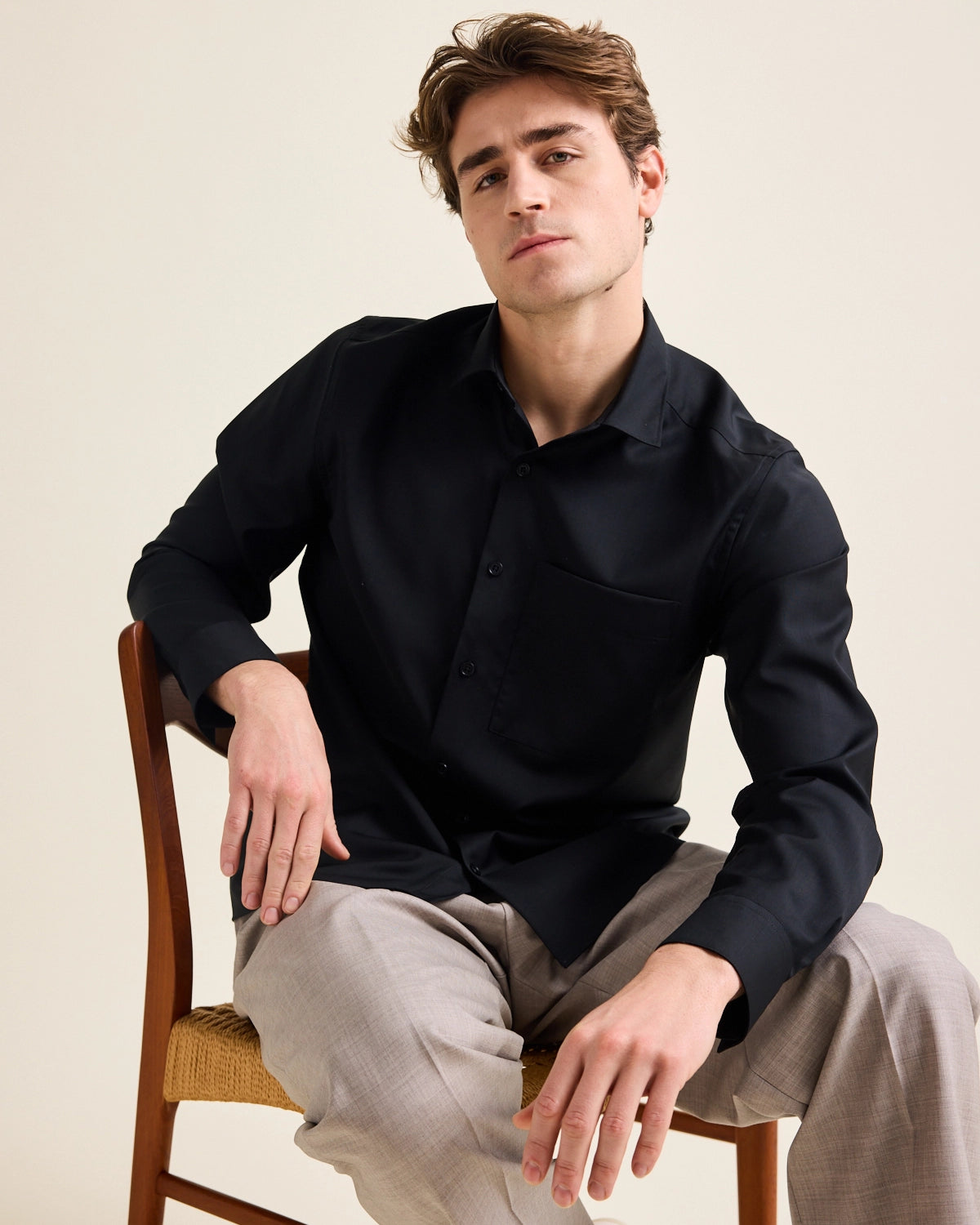 Man sits down on a vintage chair and wears a black merino wool shirt on taupe wide dressed pants.