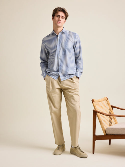 Man wears a relaxed, wide fitted merino wool shirt from Wolk in a navy-white stripe pattern. He wears it on a beige trousers.