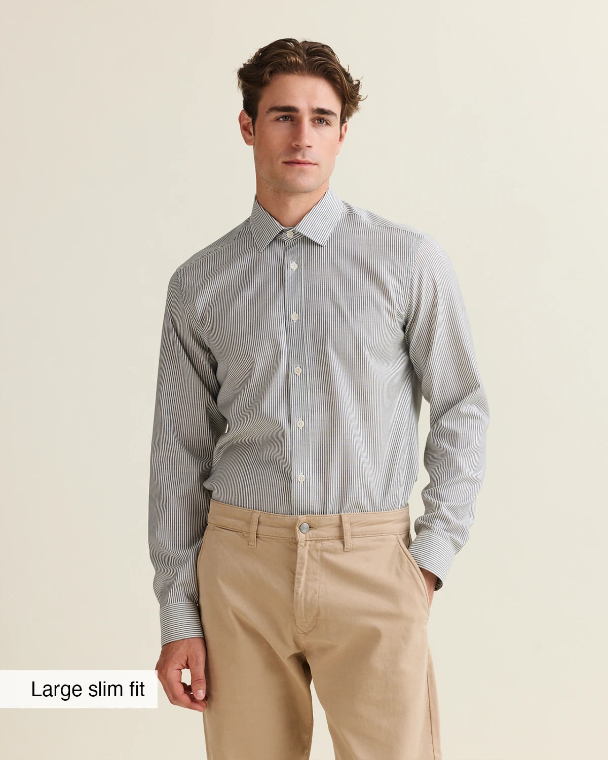 Man wearing a navy bengal striped merino shirt tucked in and beige pants on a beige background