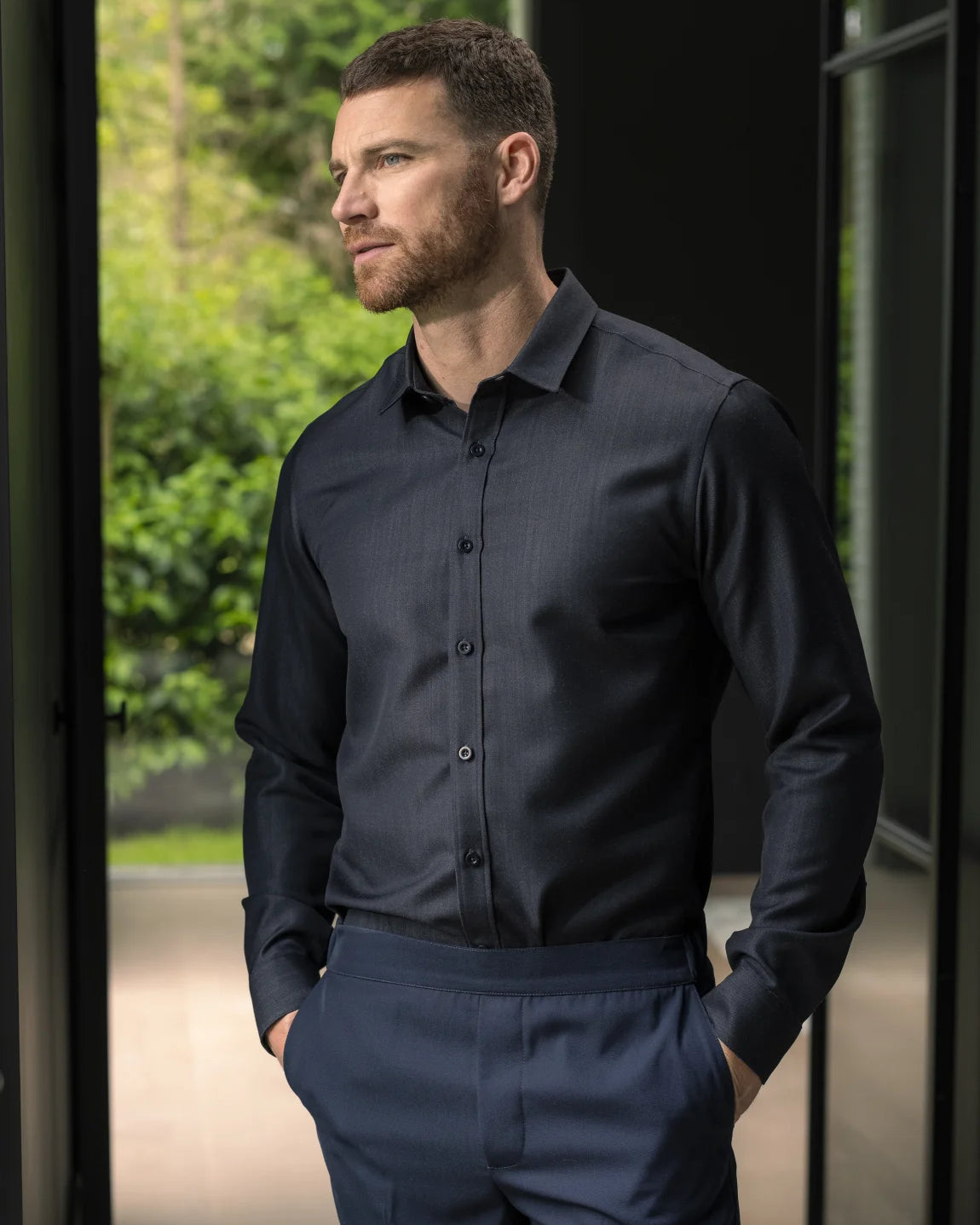 A man wearing a dark navy long-sleeve button-down merino wool denim shirt and matching navy trousers, standing in a doorway with a blurred green outdoor background.