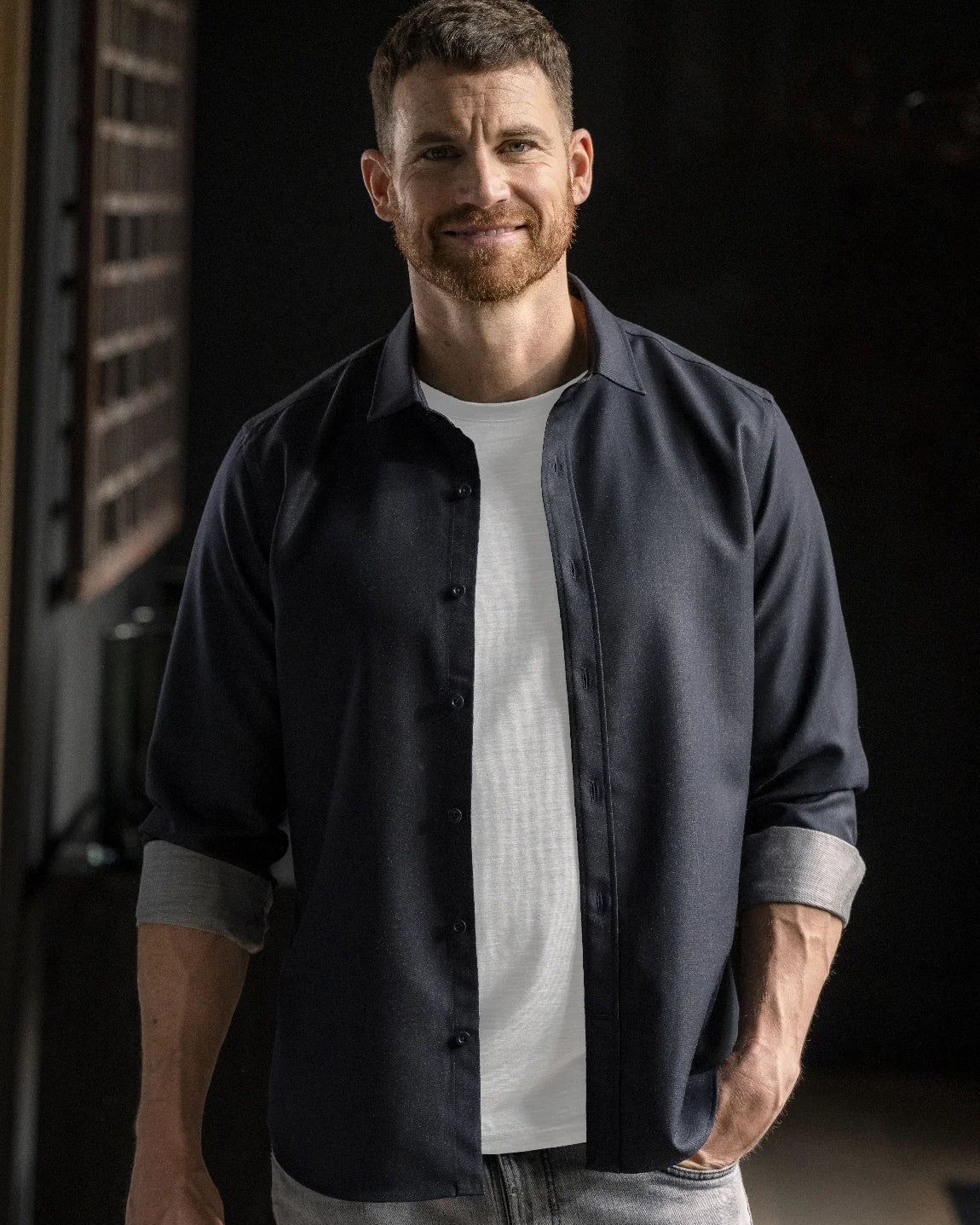 A man wearing a dark navy blue button-down shirt over a white t-shirt and light gray jeans, standing in a dimly lit indoor setting.