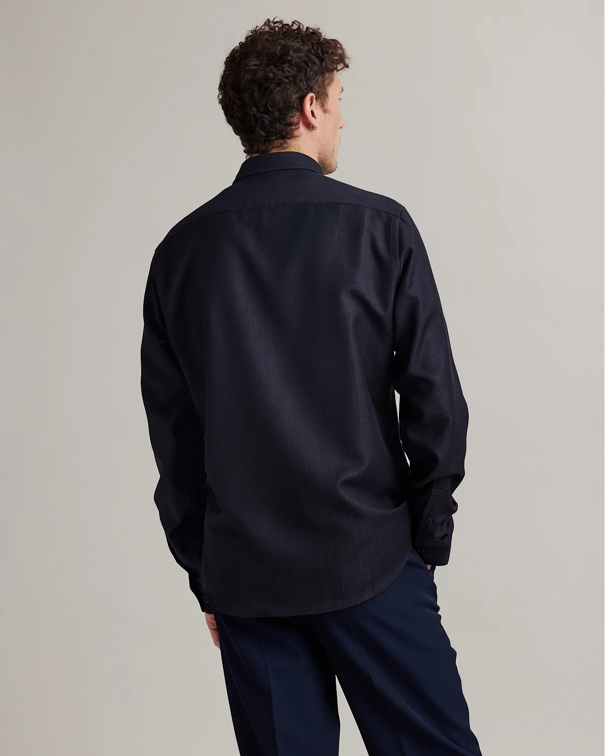 A man shown from the back wearing a dark navy blue long-sleeve button-down merino wool denim shirt and matching navy trousers, photographed against a solid light gray studio background.