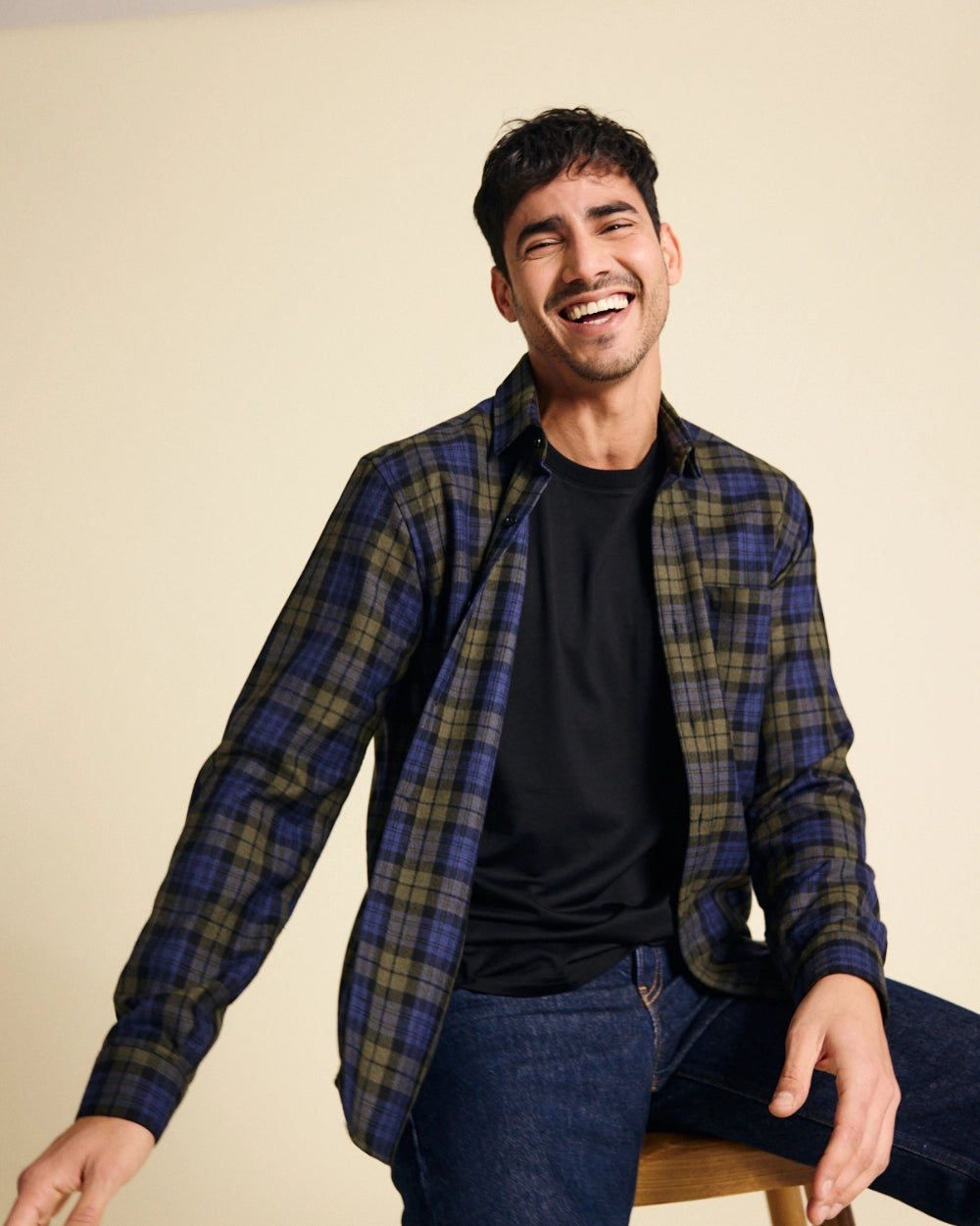 Man wearing a plaid merino flannel shirt over a black t-shirt, sitting on a stool against a beige background