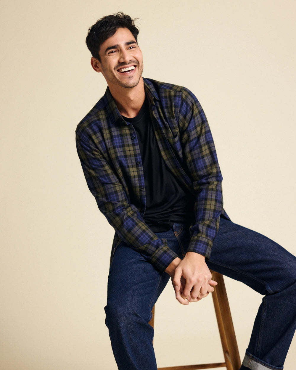 Man wearing a merino wool plaid shirt and jeans sitting on a stool against a beige background