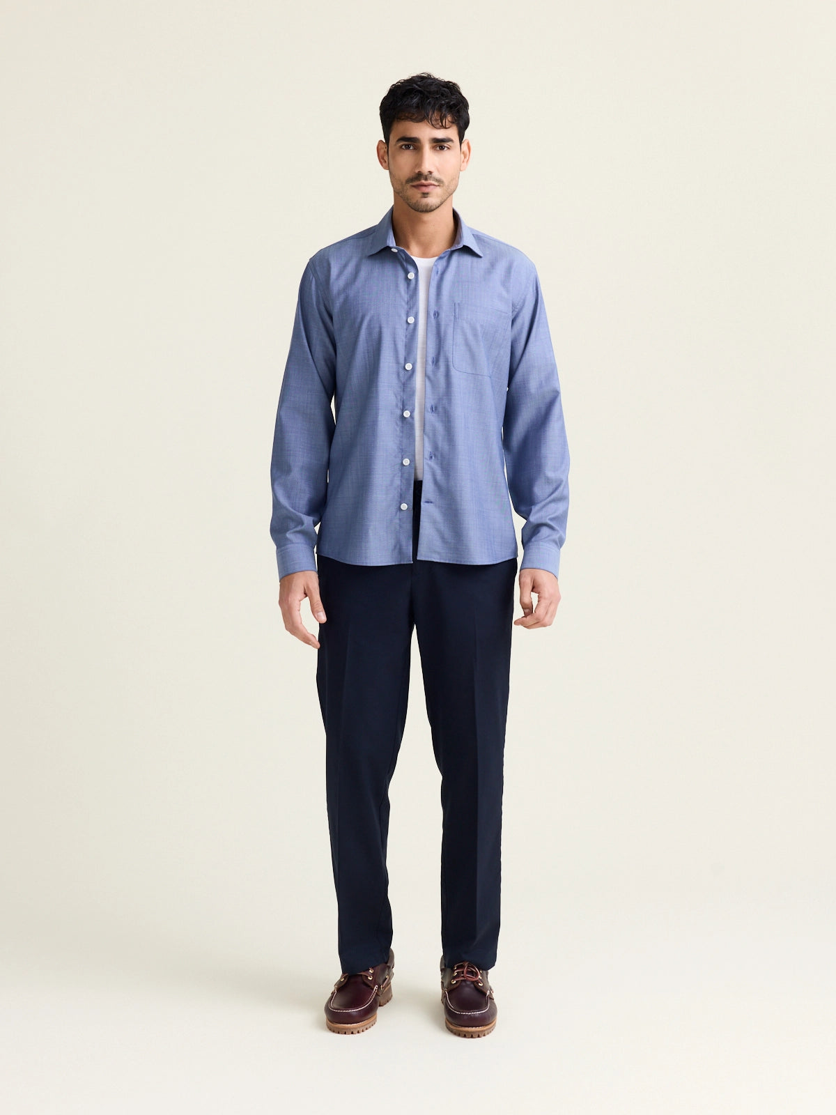 Man wears a merino wool shirt in light blue denim color with chest pocket and white buttons open on a white merino wool T-shirt and on a dark navy pants
