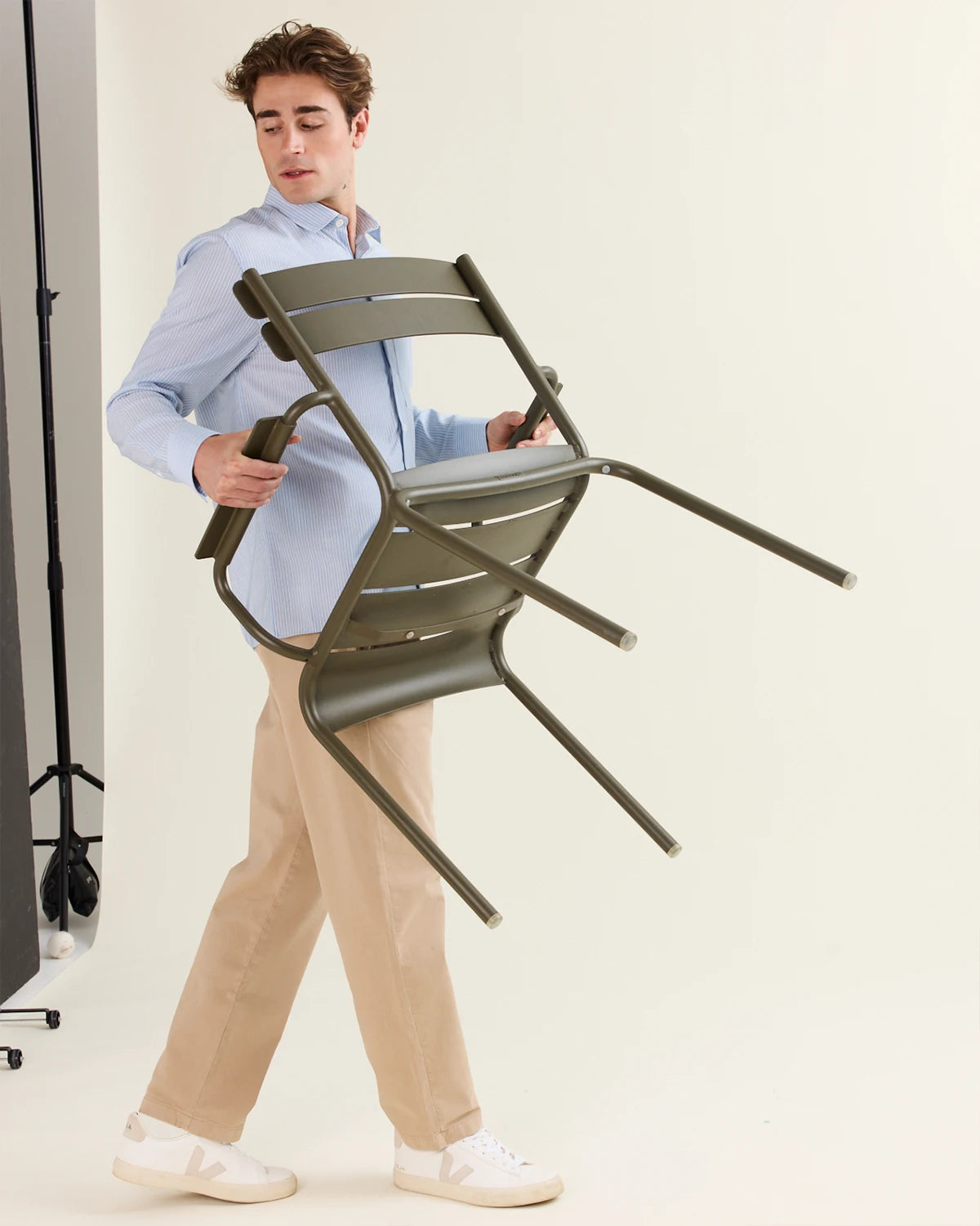Man walking on a photoshoot set holding a green chair and wearing a light blue striped merino shirt on a beige trousers