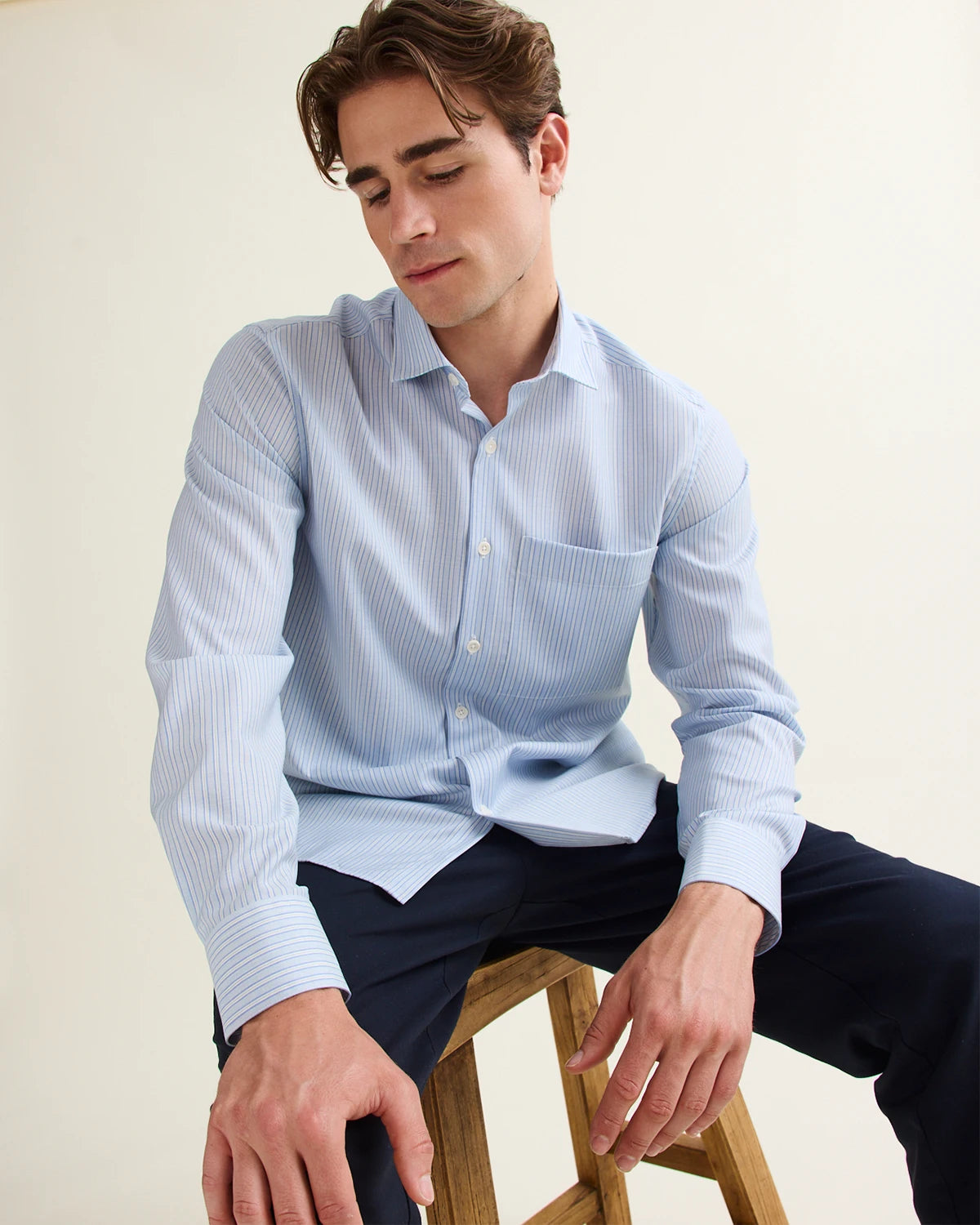 Man wearing a light blue striped merino wool shirt with square chest pocket sitting on a wooden stool against a plain background