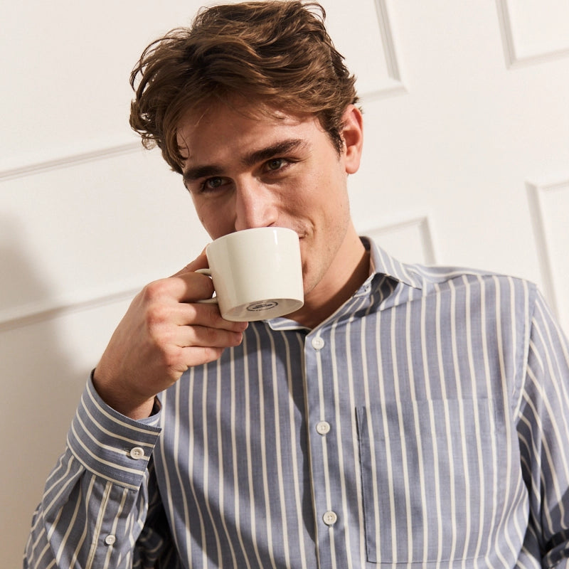 Man drinking from a white mug against a light background wearing a navy stripe merino wool shirts with long sleeves and a chest pocket