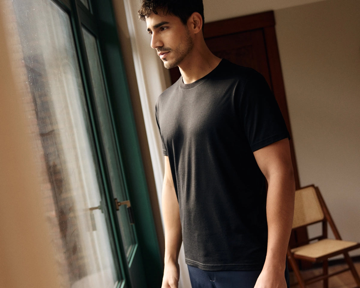 Man wearing a black merino wool T-shirt and is standing by a window in a room with a chair in the background
