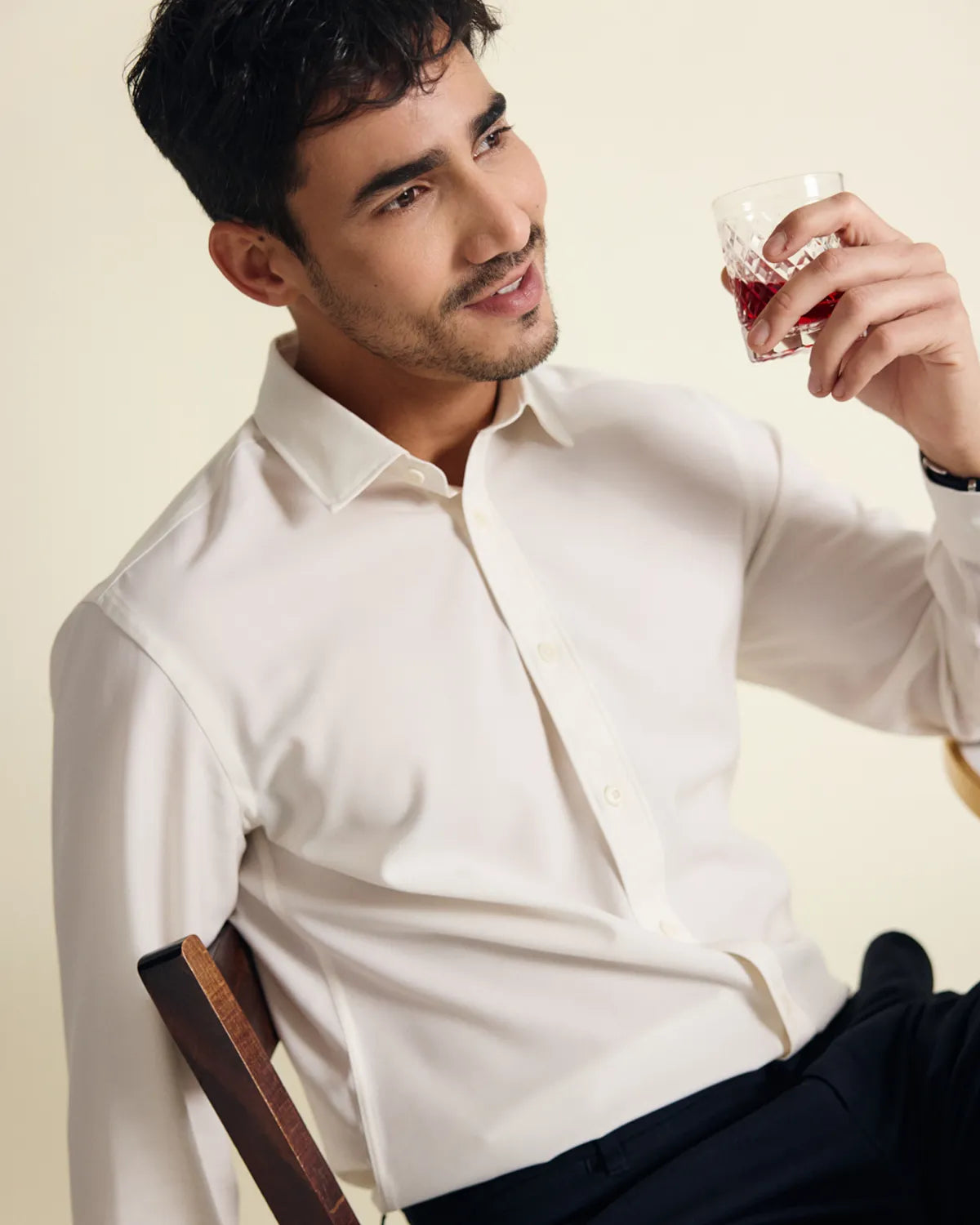 Man in a white merino dress shirt sits on a wooden chair with a cocktail against a beige background