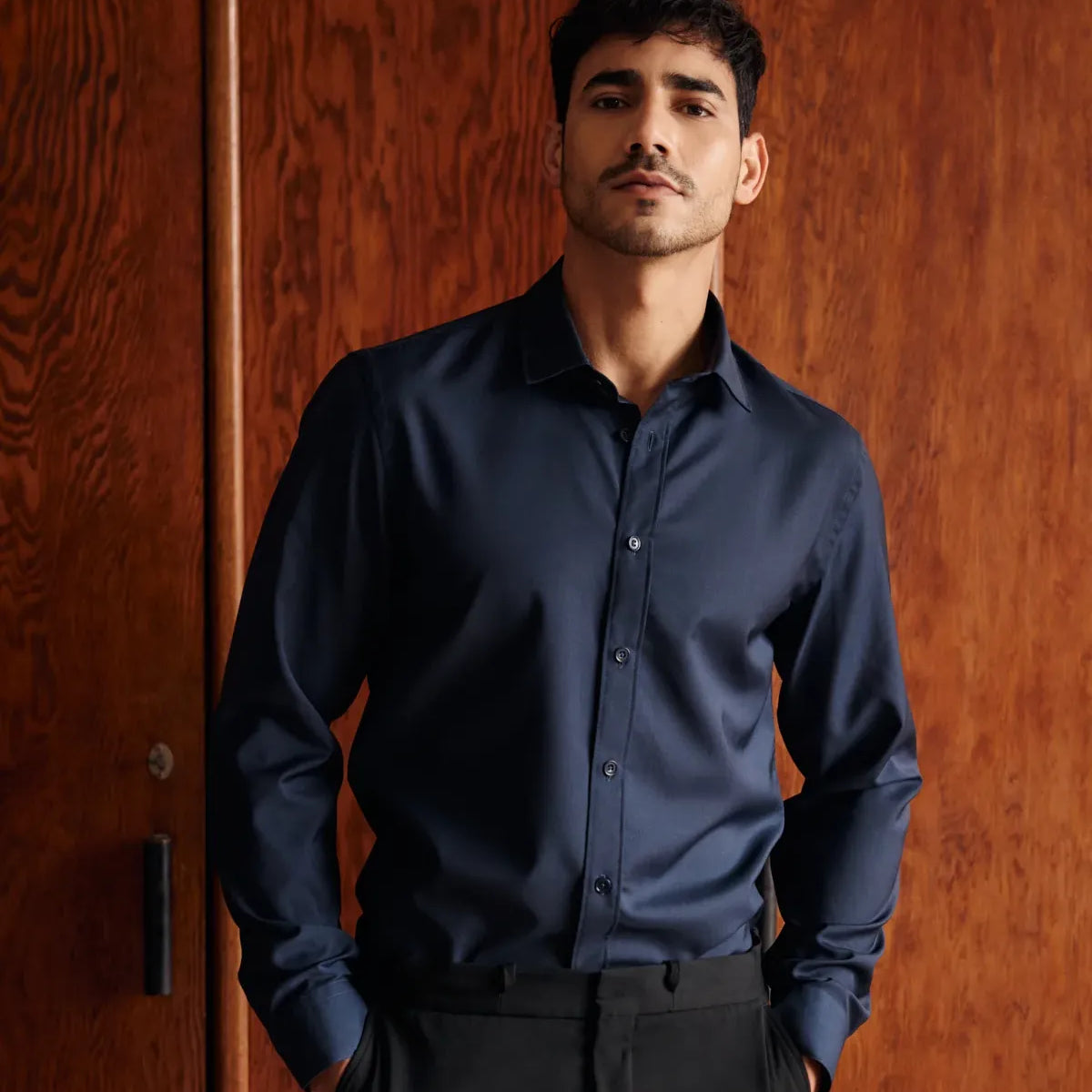 Man wearing a navy blue merinowool dress shirt in slim fit and black pants against a wooden door background