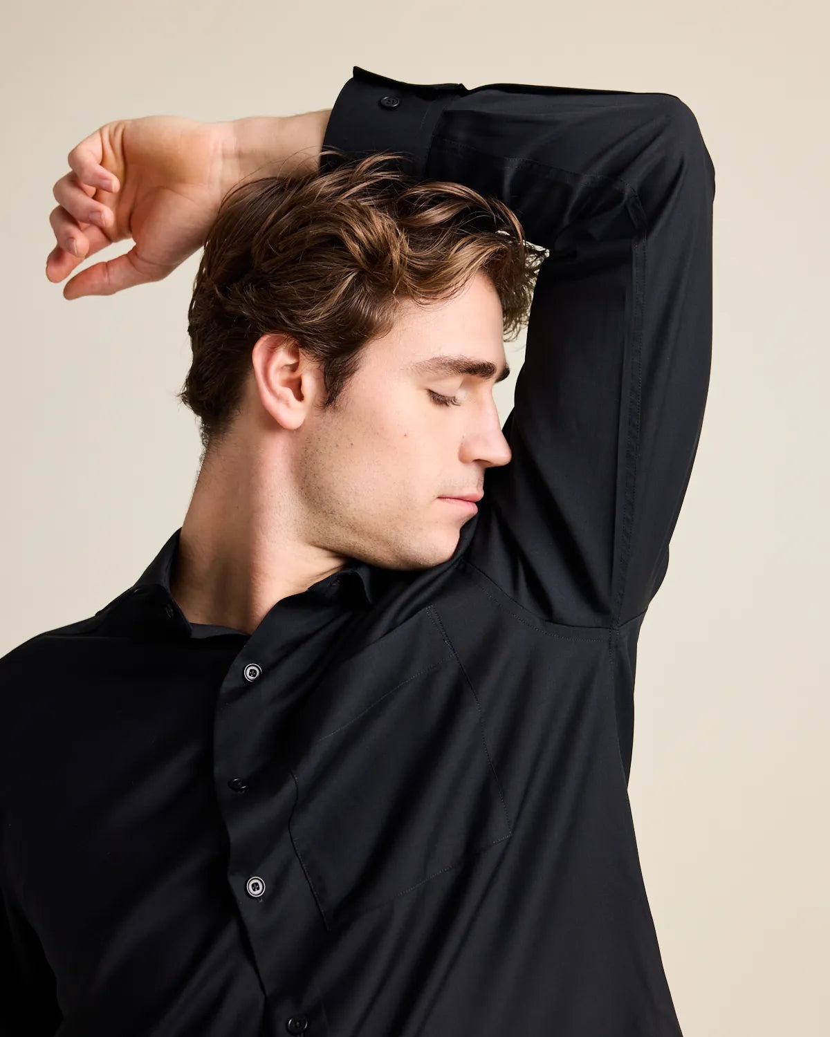 Man smelling the fresh armpit of a black merino shirt with one arm raised against a beige background