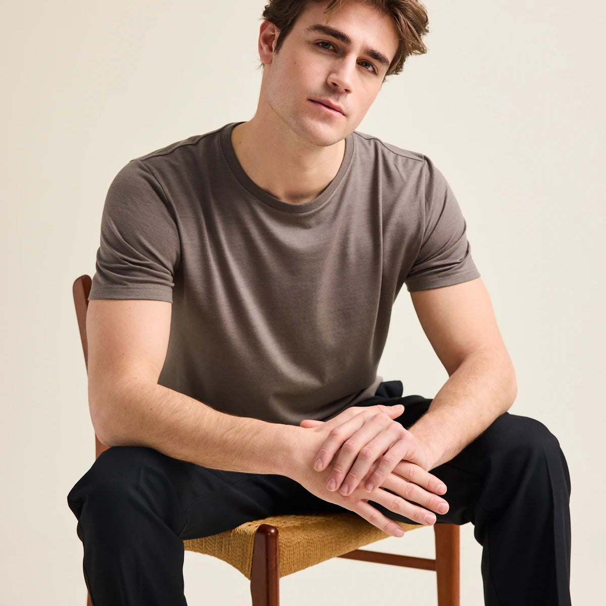 Man sitting on a chair wearing a brown merino t-shirt from Wolk and black pants against a beige background