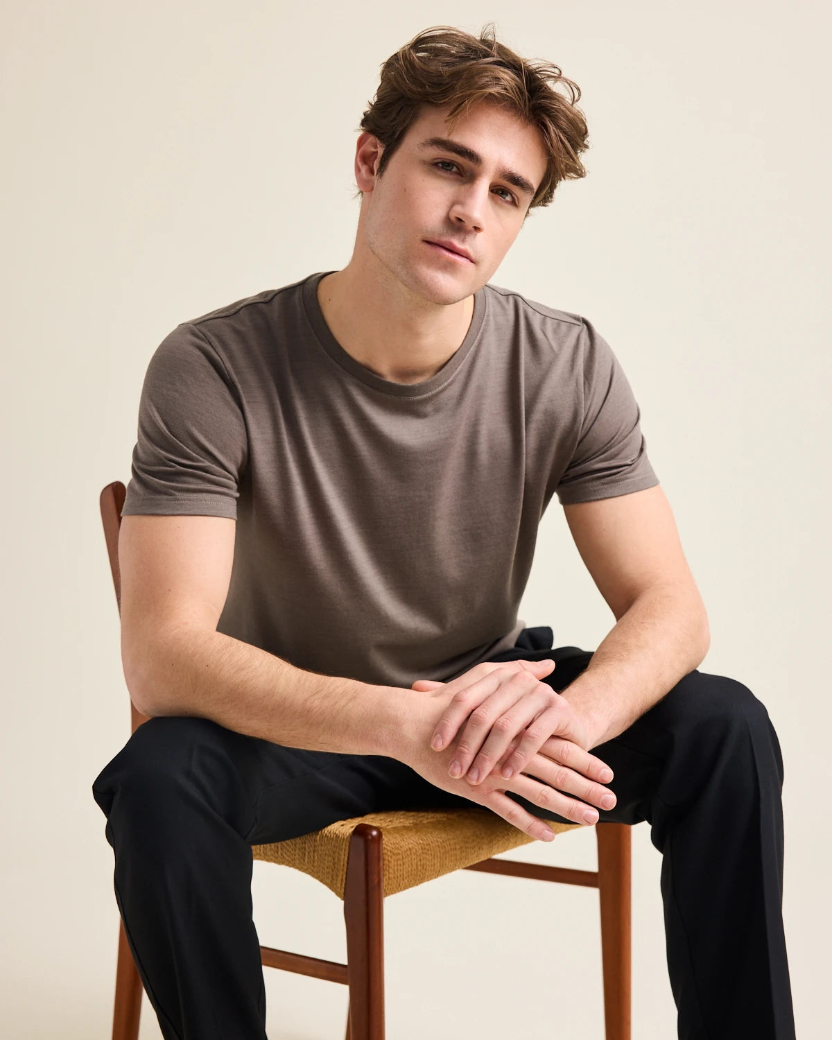 Man sitting on a chair wearing a brown merino t-shirt from Wolk and black pants against a beige background