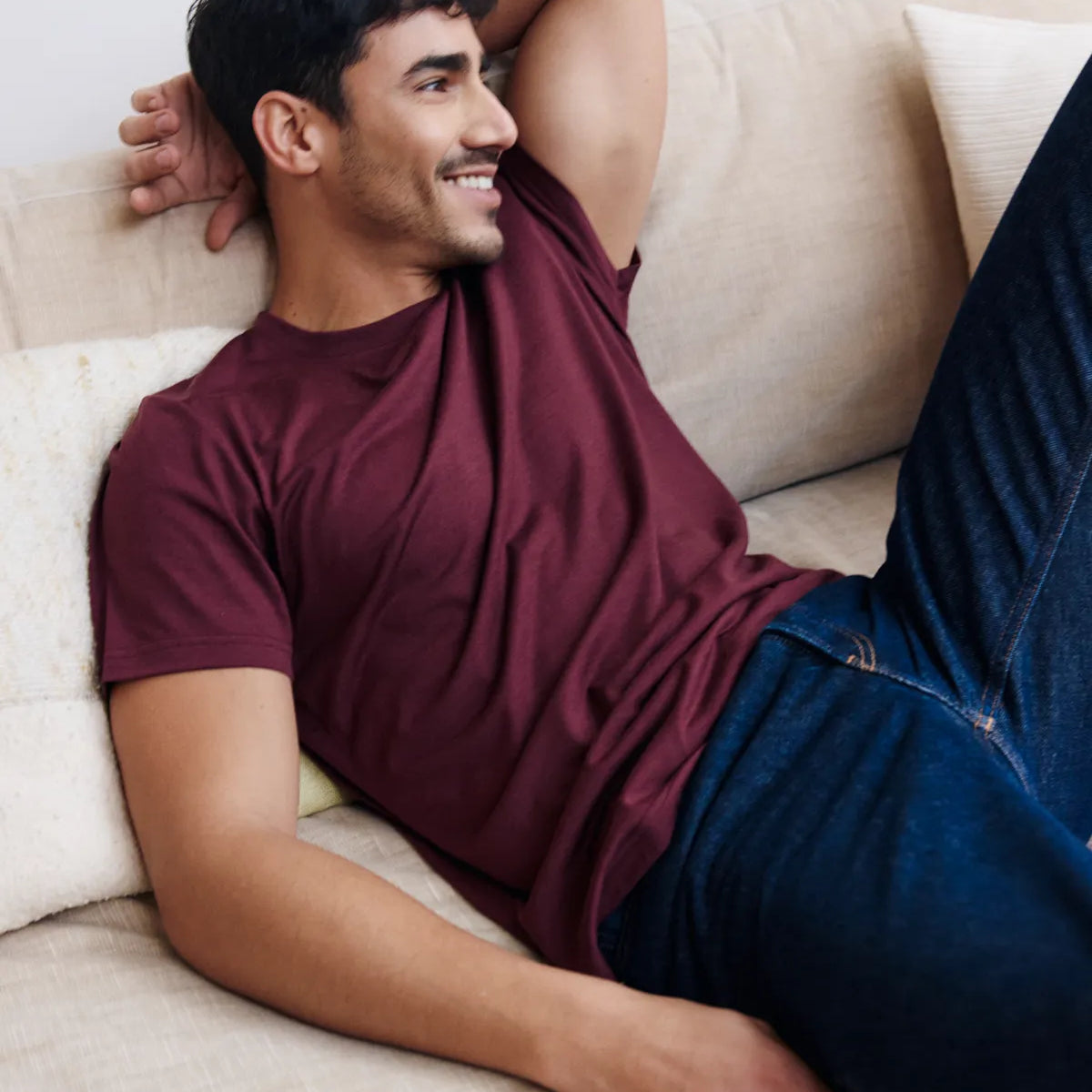 Man in a burgundy merino wool t-shirt from Wolk and blue jeans lying on a beige couch.