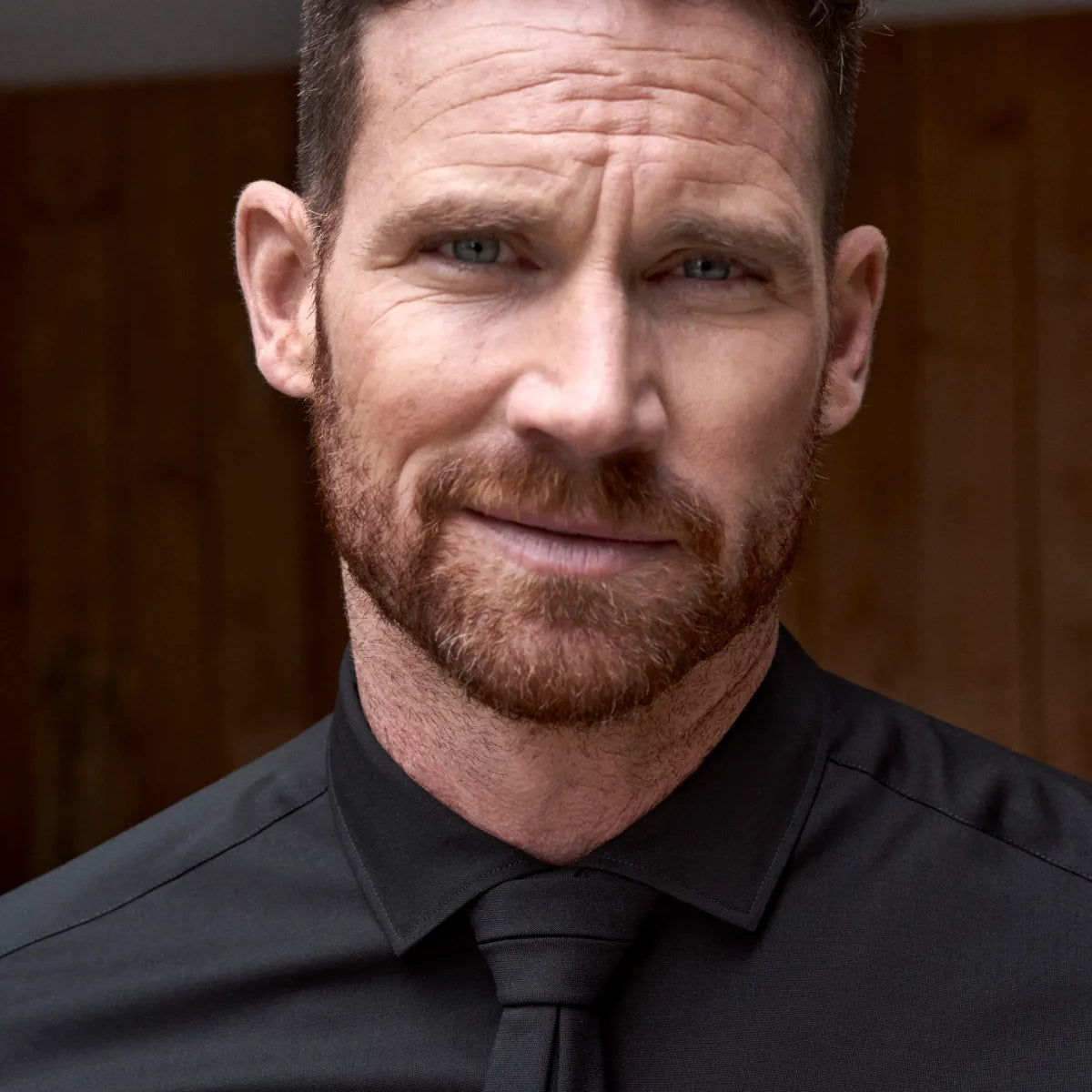 Man wearing a black merinowool shirt and a black tie against a wooden background