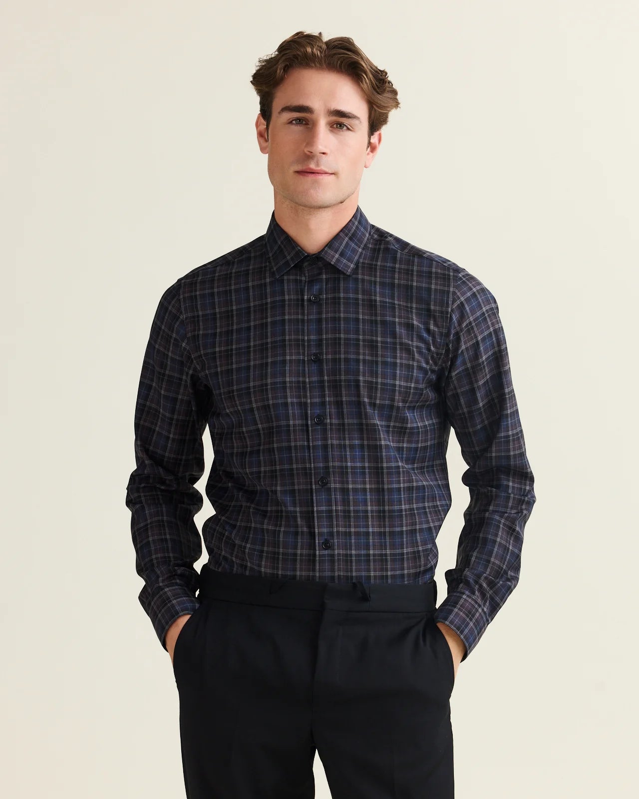 A man wearing a long-sleeve button-up plaid merino shirt in navy, purple and blue with thin grey and black lines, paired with dark trousers, standing against a light beige background.
