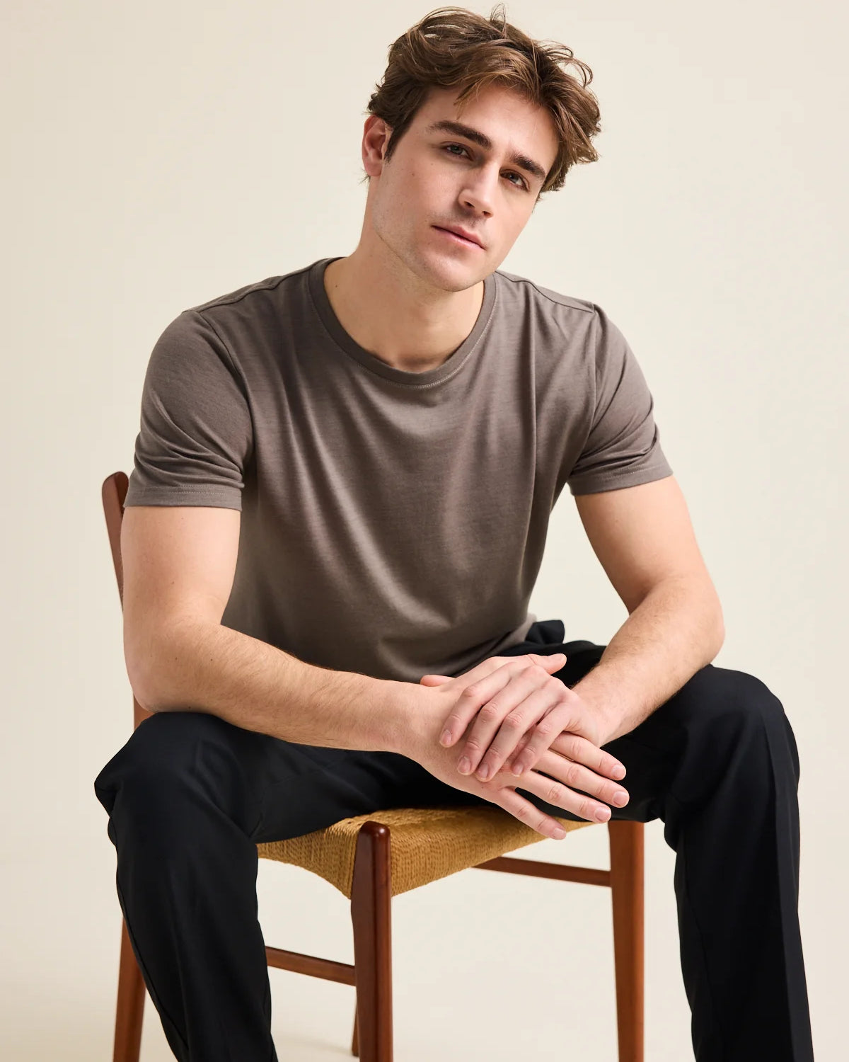 A male model is wearing a chocolate brown merino wool T-shirt from the brand Wolk while he sits on a chair in a studio setting. The T-shirt has a crew neck.