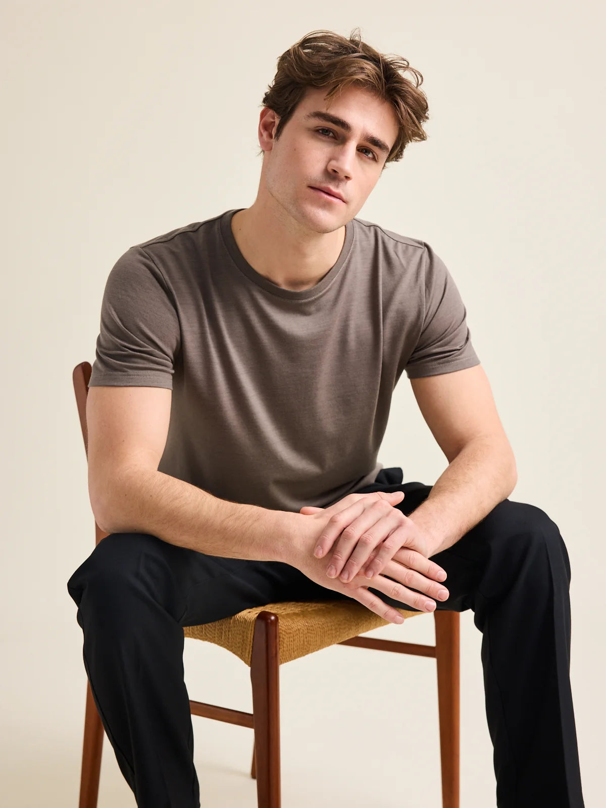 A male model is wearing a chocolate brown merino wool T-shirt from the brand Wolk while he sits on a chair in a studio setting. The T-shirt has a crew neck.