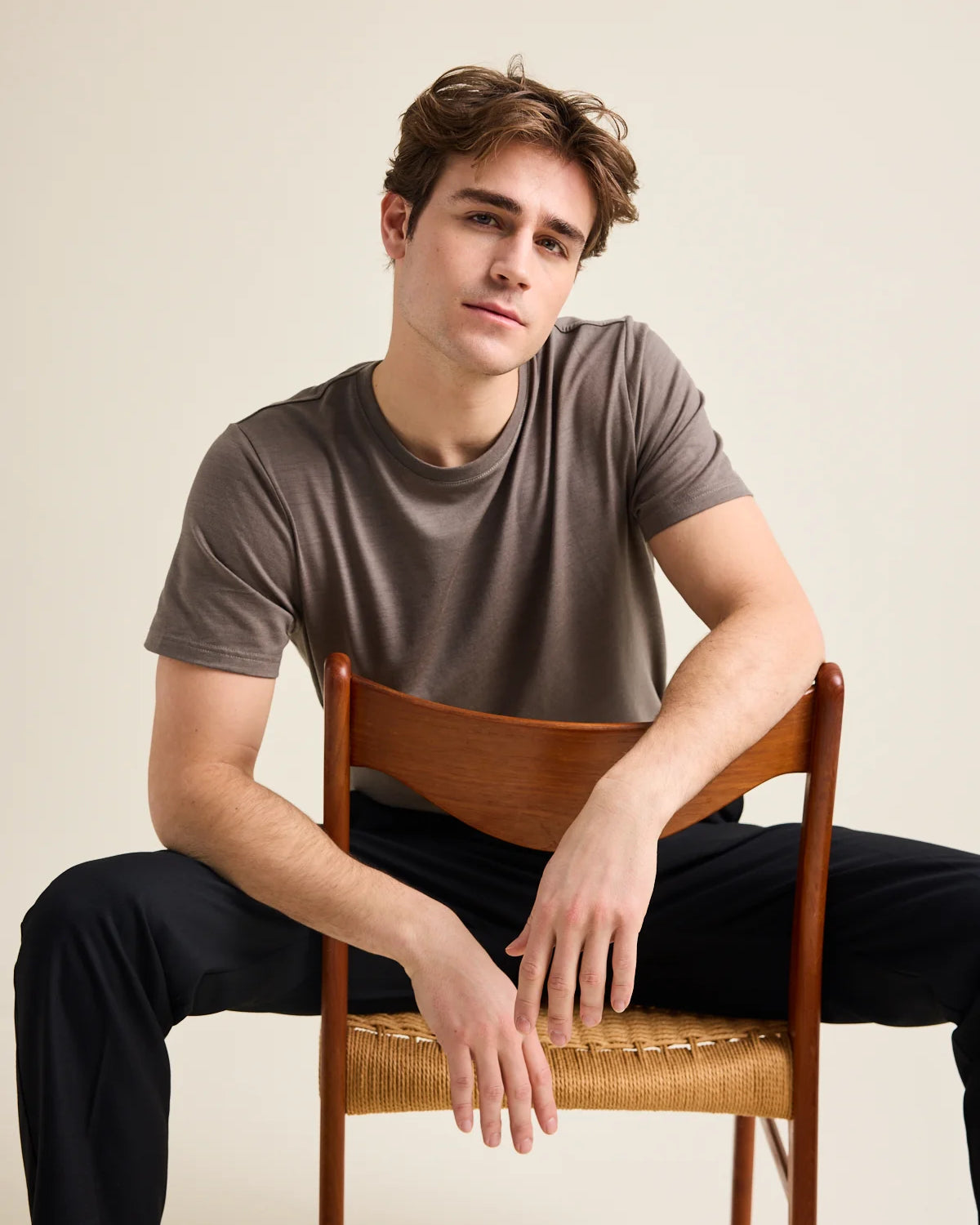 A male model, wearing a chocolate brown merino wool T-shirt from the brand Wolk. He sits on a vintage chair and leans on the back-rest.