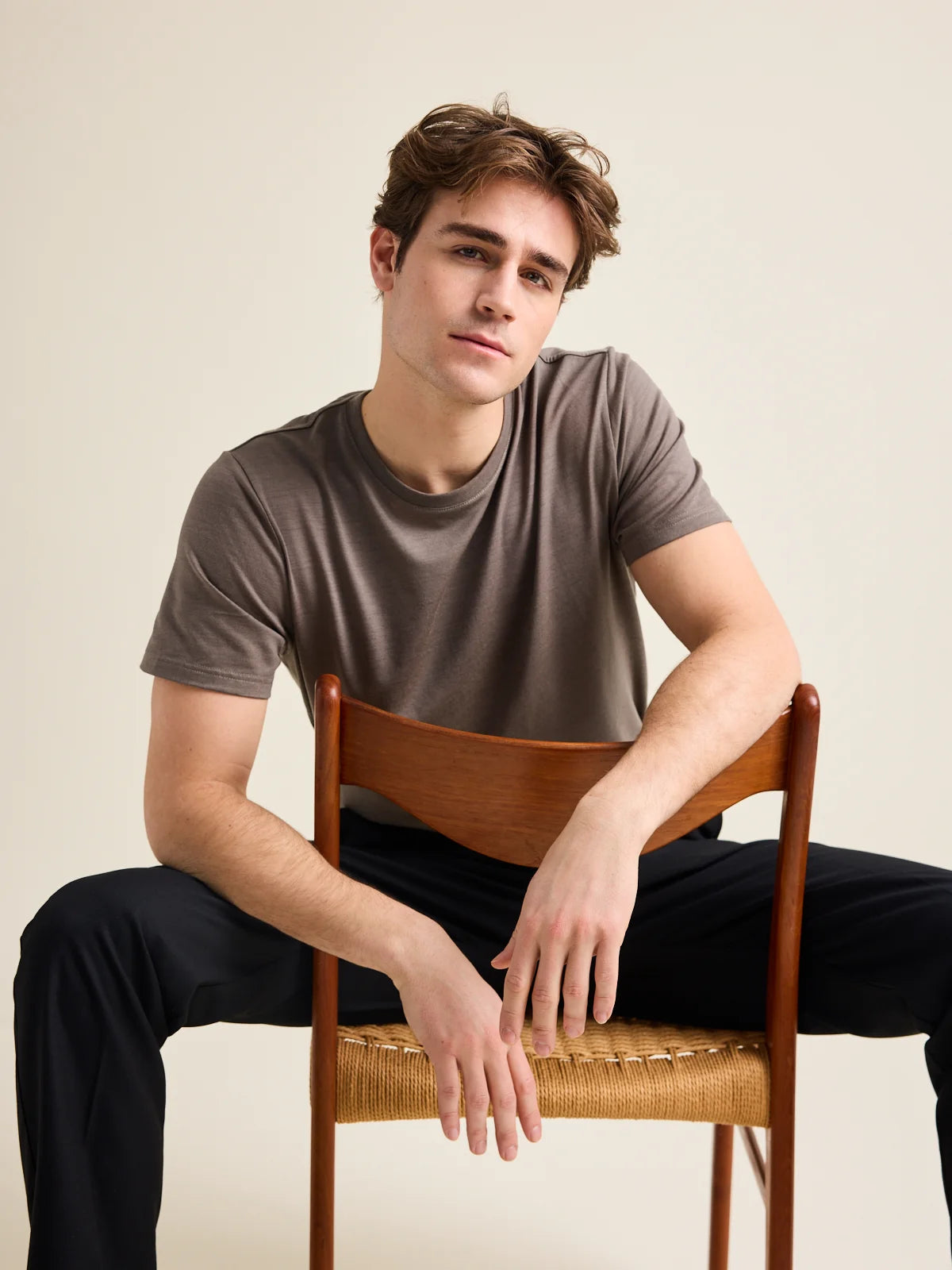 A male model, wearing a chocolate brown merino wool T-shirt from the brand Wolk. He sits on a vintage chair and leans on the back-rest.