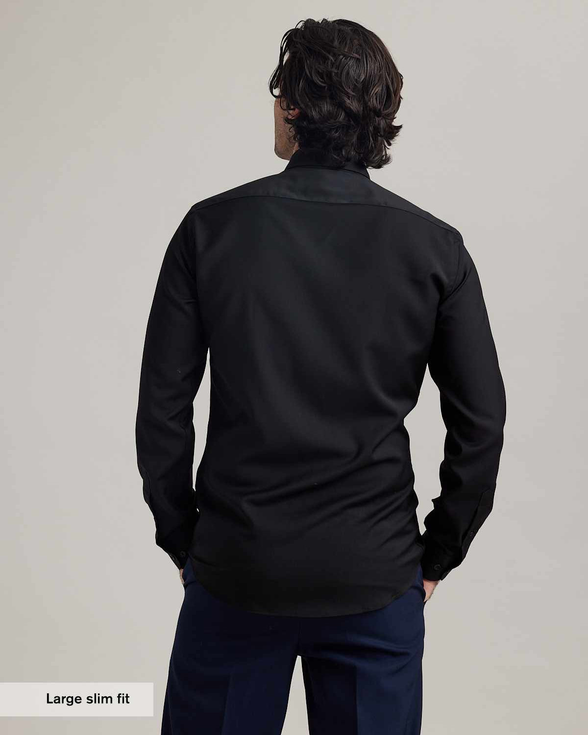 back view of a man wearing a black merino dress shirt and blue pants on a plain background. It shows the back yoke and the shirt length.