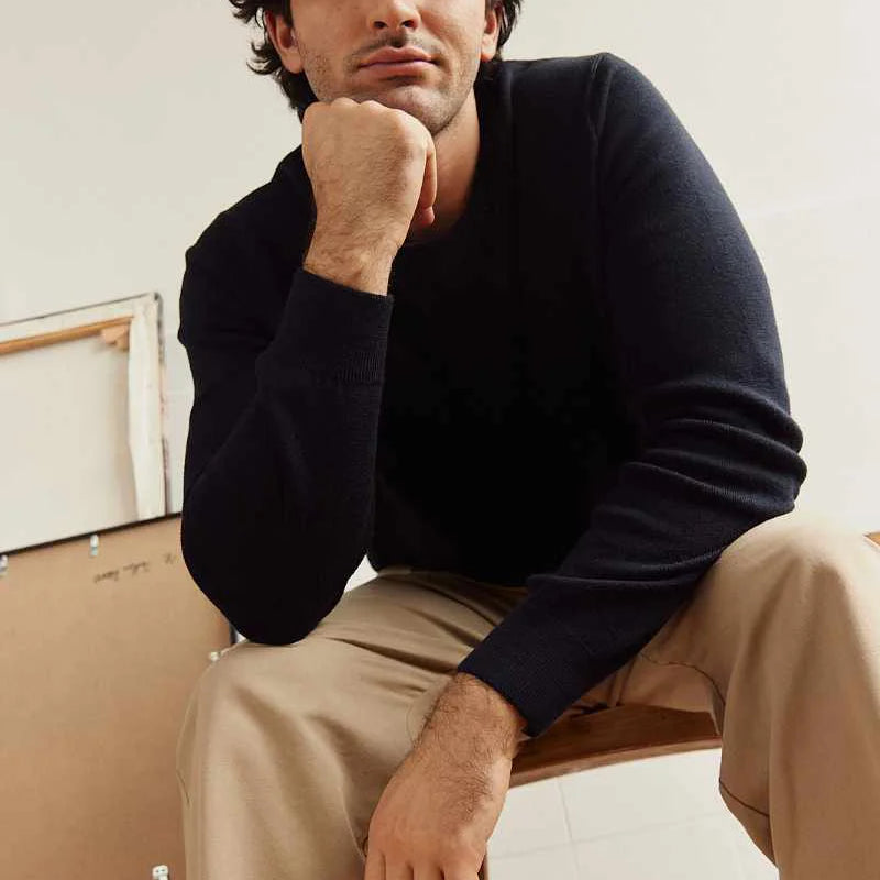 Man sitting on a chair wearing a dark navy merino wool sweater and beige pants against a neutral background