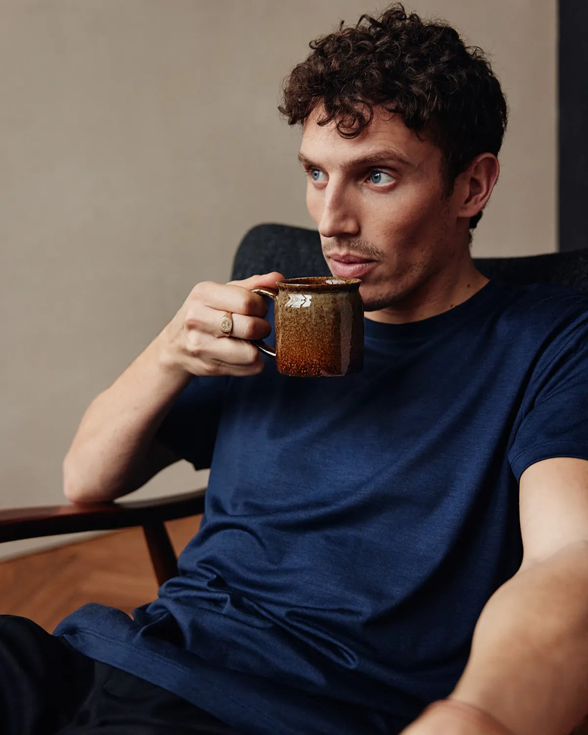 man wearing a navy coloured merino T-shirt from Wolk, drinking a coffee while he sits in an armchair