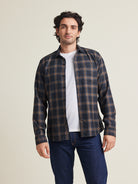 Man wears a merino wool flannel shirt in dark grey brown plaid with chestpocket open over a white merino wool T-shirt on a dark denim jeans