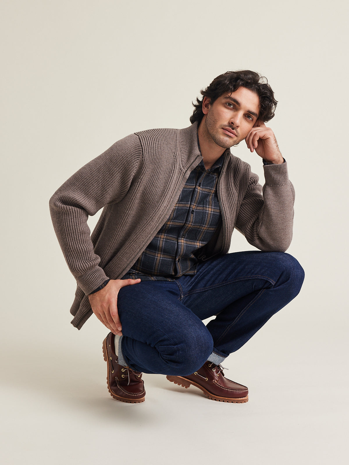 Man wears a merino wool vest in brown on a merino wool flannel shirt in grey brown plaid pattern with chest pocket on dark blue denim with bootshoes.