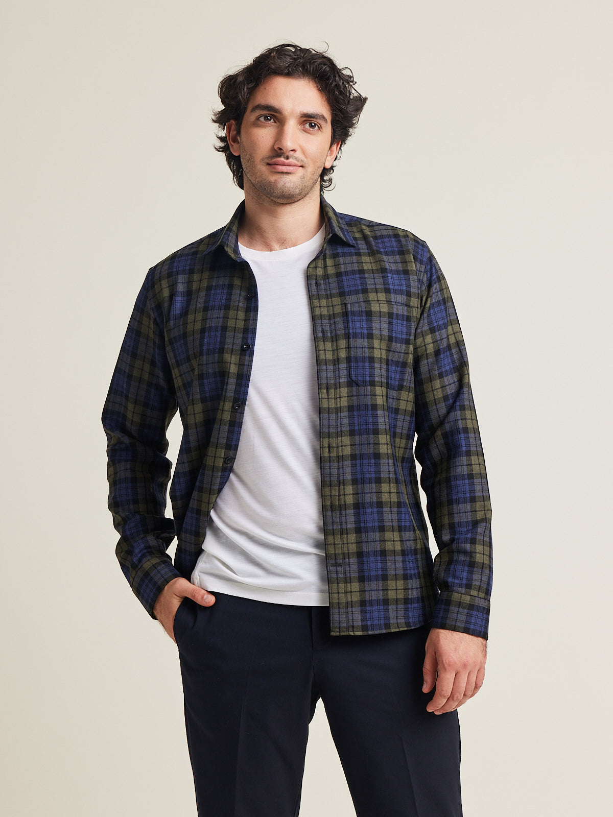 Man wears a merino flannel shirt in plaid blue green color with chestpocket open over a white merino wool T-shirt on a dark navy pants