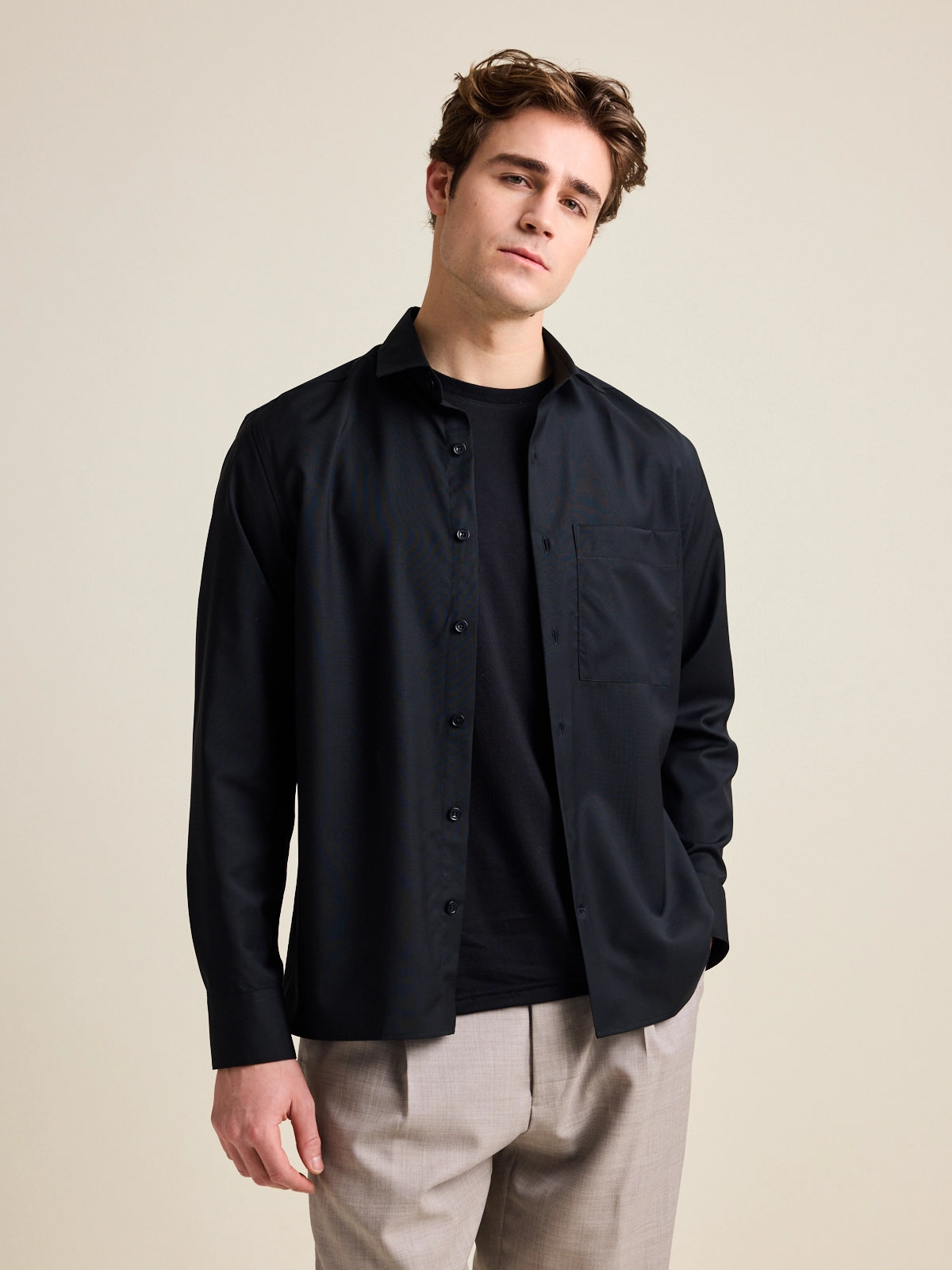 Man wears a black merino wool shirt open over a black merino wool T-shirt from Wolk. The shirt has a relaxed, wide fit.