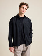 Man wears a black merino wool shirt open over a black merino wool T-shirt from Wolk. The shirt has a relaxed, wide fit.