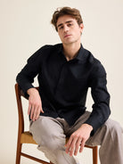 Man sits down on a vintage chair and wears a black merino wool shirt on taupe wide dressed pants.