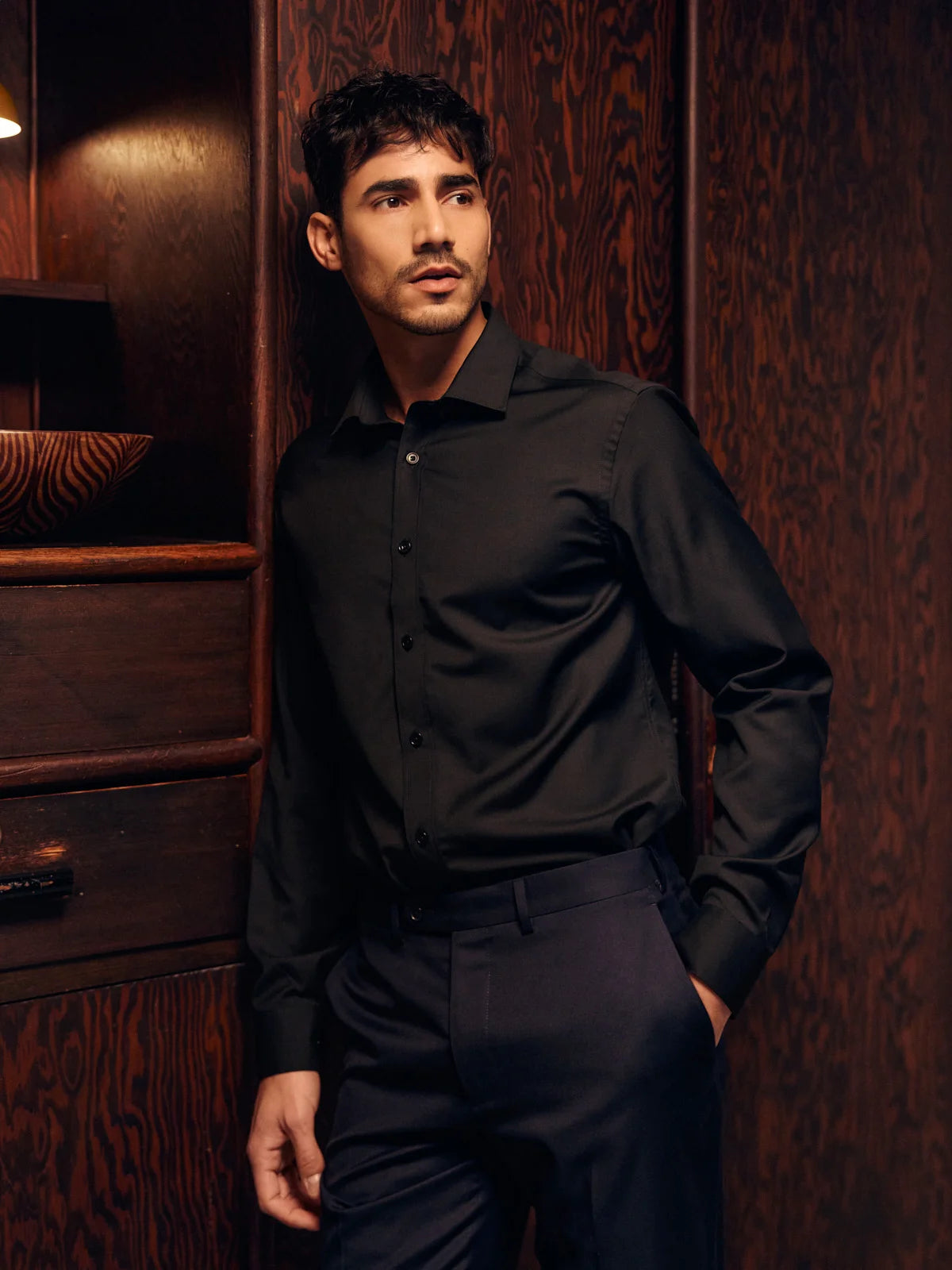 Man wearing a black merino wool shirt and pants standing against a wooden wall.