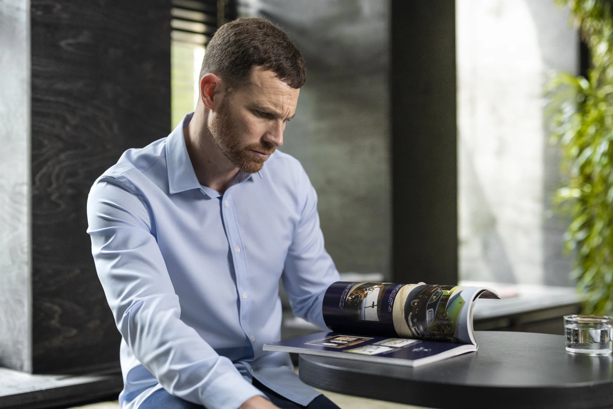 Man reading a magazine at a table with a blurred background wearing a light blue merino wool shirt from wolk