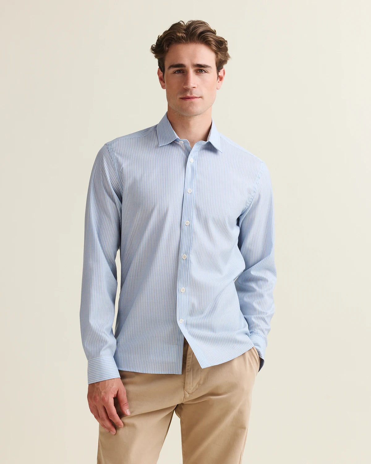 Man wearing a light blue striped merino shirt and beige pants against a beige background