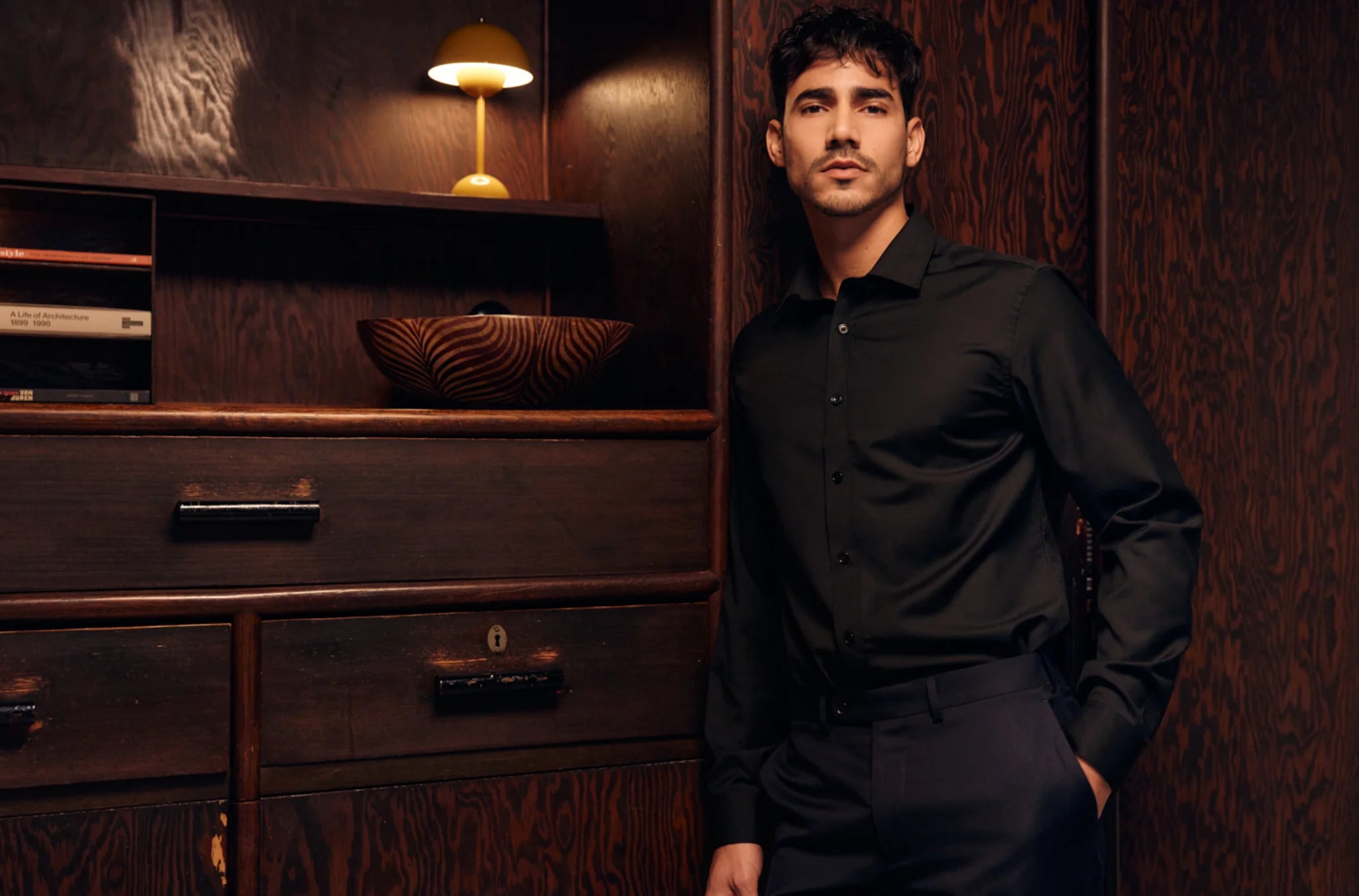 Man in a black merino wool shirt standing in a room with wooden furniture and decor.