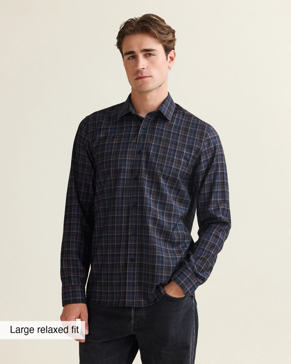 A man wearing a dark navy and black plaid long-sleeve button-up merino wool shirt with a relaxed fit, paired with dark jeans, against a light beige background.