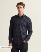 A man wearing a dark navy and black plaid long-sleeve button-up merino wool shirt with a relaxed fit, paired with dark jeans, against a light beige background.