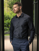 A man wearing a dark navy long-sleeve button-down merino wool denim shirt and matching navy trousers, standing in a doorway with a blurred green outdoor background.