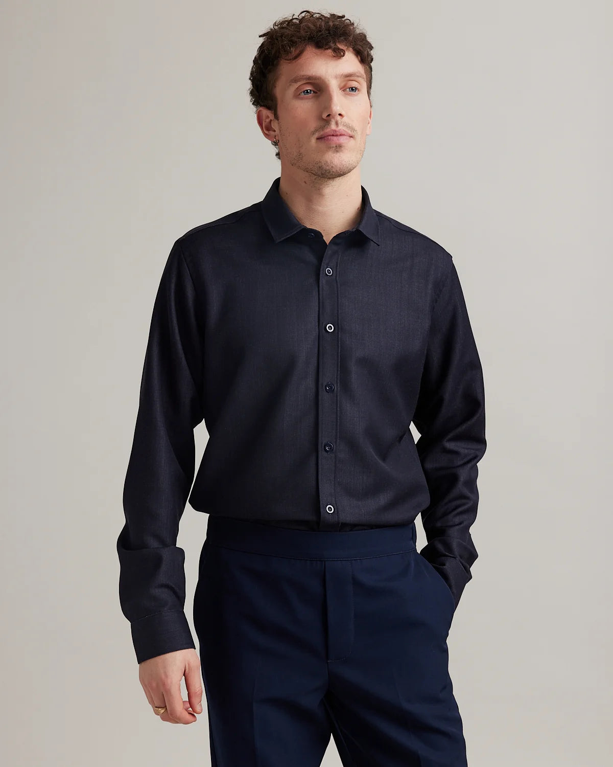A man wearing a dark navy blue long-sleeve button-down shirt in merino wool denim with matching navy trousers, standing against a neutral light gray studio background.