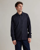 A man wearing a dark navy long-sleeve button-up shirt made from merino wool denim and matching navy trousers, standing against a plain light gray studio background.