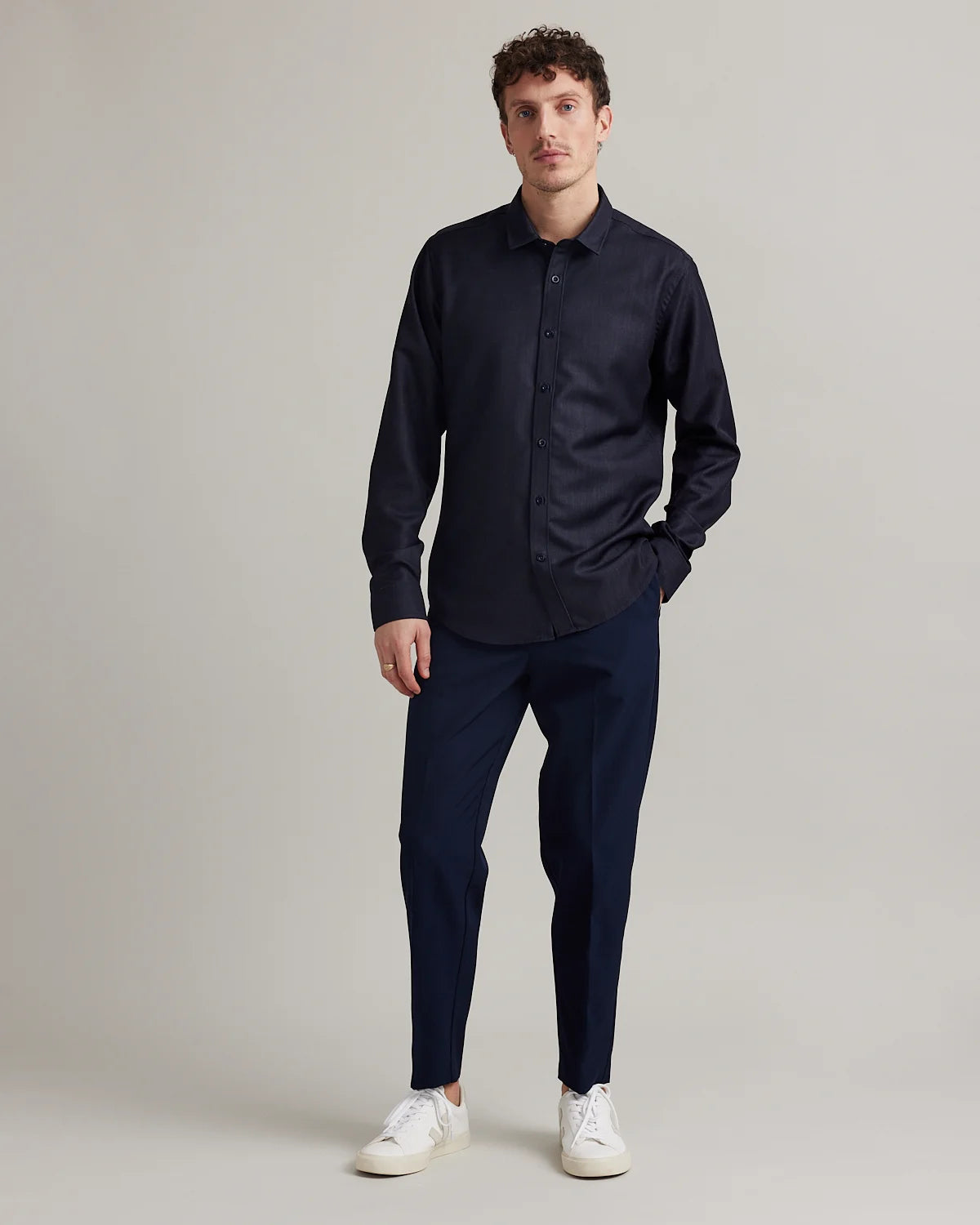 A man modeling a dark navy blue long-sleeve button-down shirt and matching navy trousers, paired with white sneakers, against a neutral light gray studio background.
