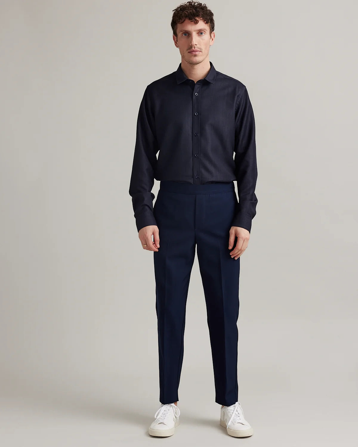 A man modeling a dark navy blue long-sleeve button-down merino wool shirt and matching navy trousers, paired with white sneakers, against a neutral light gray studio background.