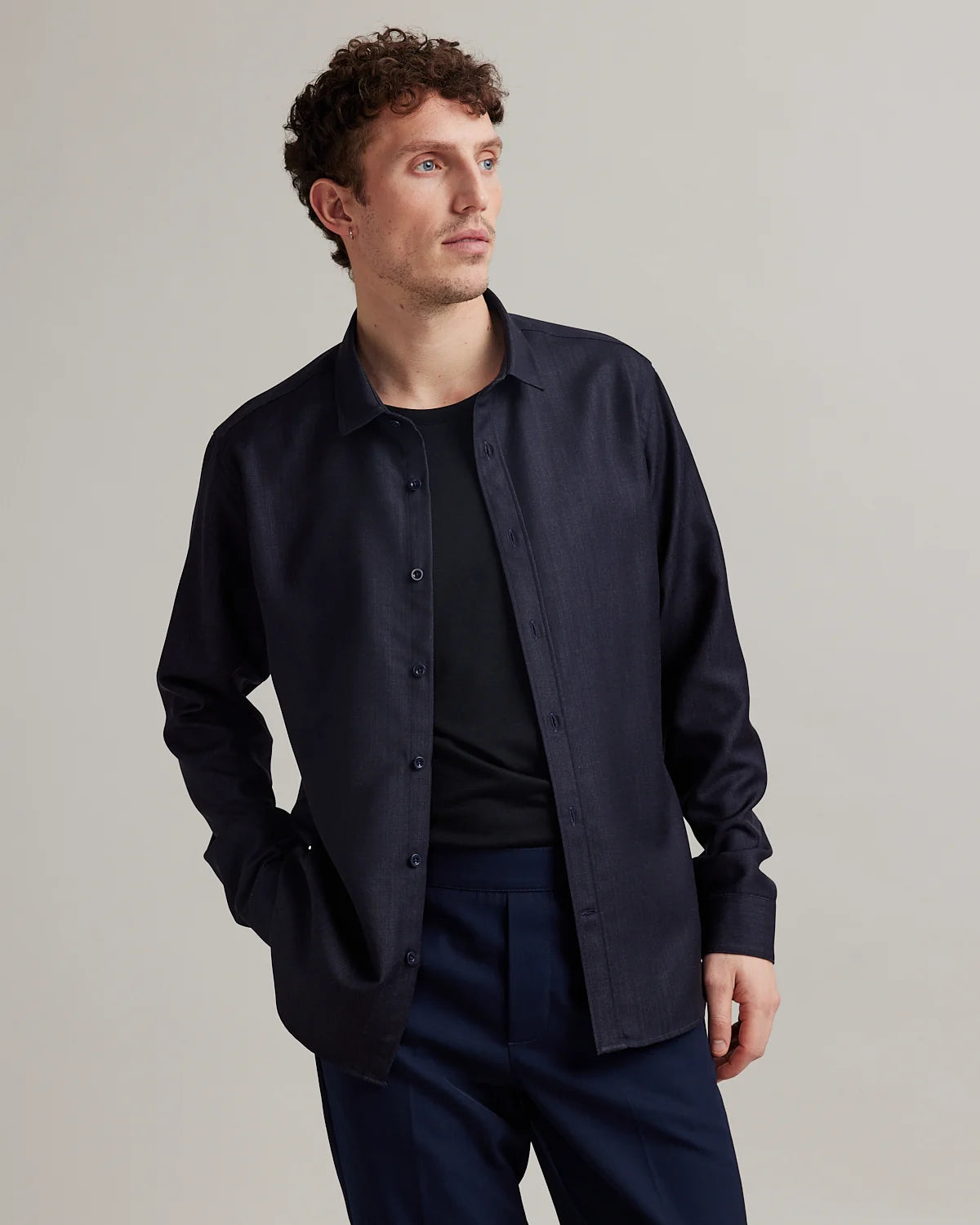 A man wearing a dark navy blue long-sleeve button-up merino wool shirt over a black merino wool t-shirt from the brand Wolk with navy trousers, photographed against a neutral light gray studio background.