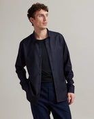 A man wearing a dark navy blue long-sleeve button-up merino wool shirt over a black merino wool t-shirt from the brand Wolk with navy trousers, photographed against a neutral light gray studio background.